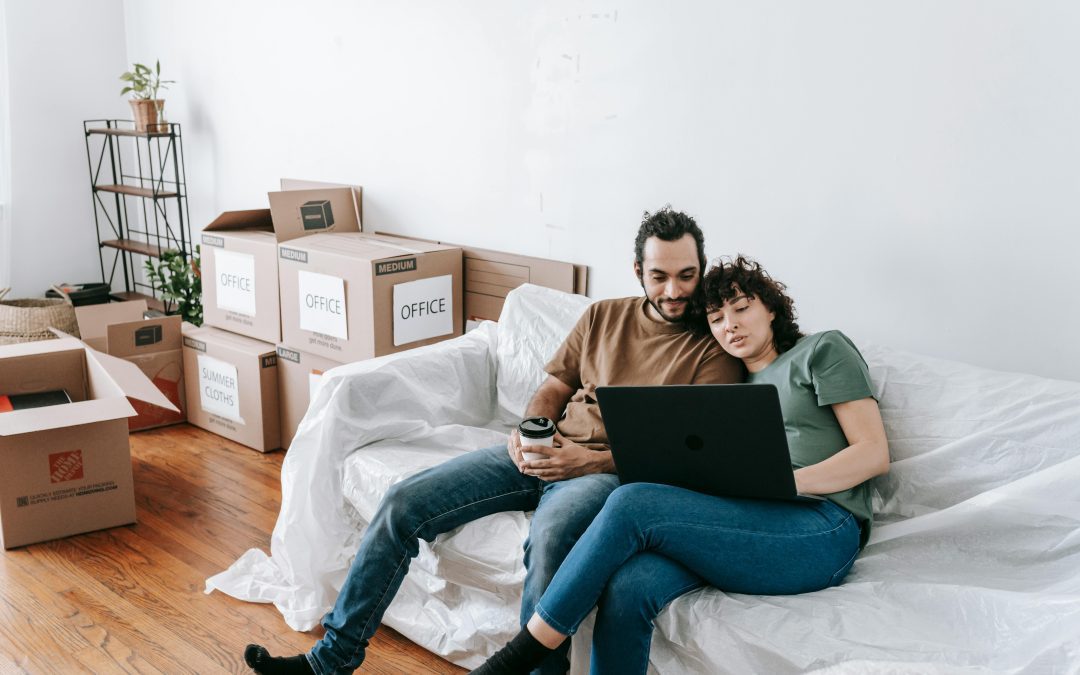 A couple sits together on a couch, using a laptop amidst moving boxes and discussing things to consider when moving in together